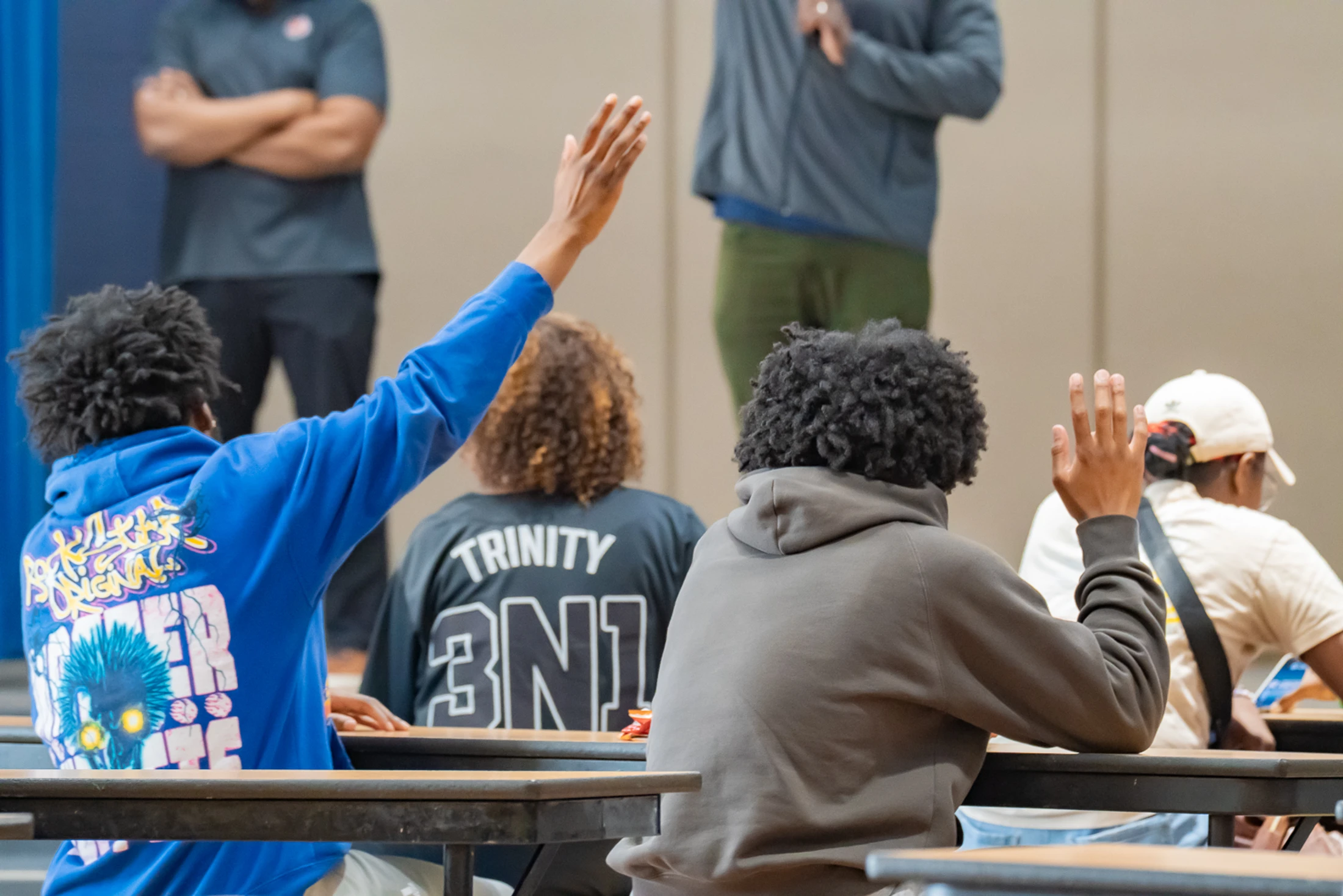 A view from behind of several students sitting at desks. Two young men have their hands raised, ready to participate in a discussion or ask a question.