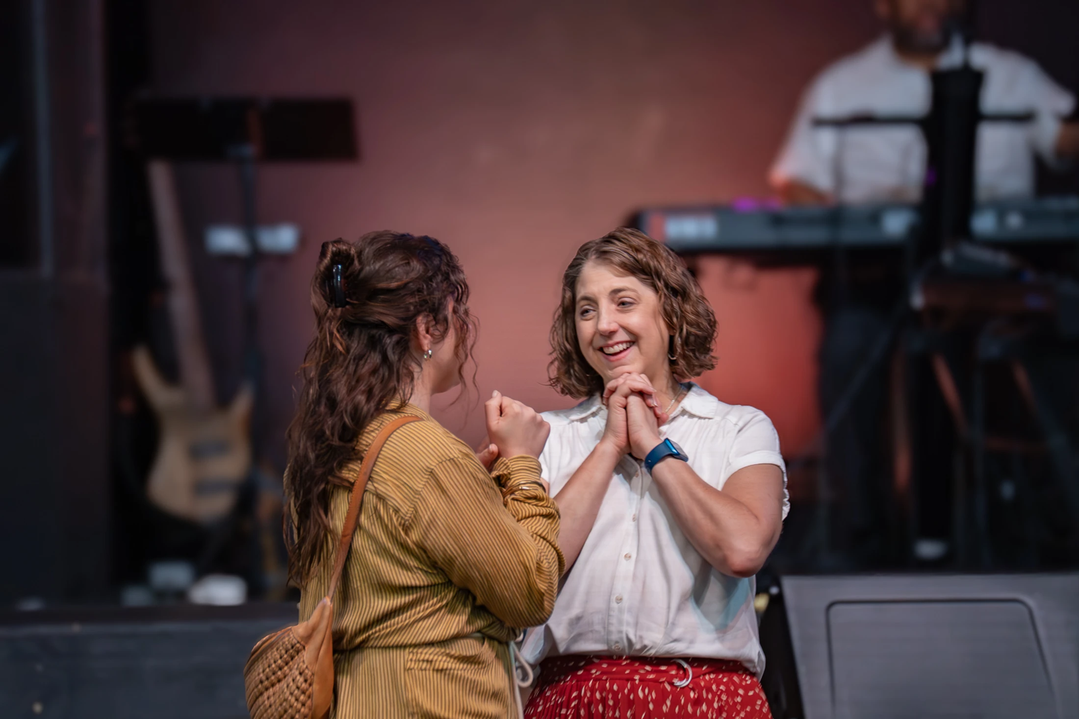 Two women stand face-to-face on a dimly lit stage. One woman smiles joyfully while holding the other's hands during an engaging conversation.