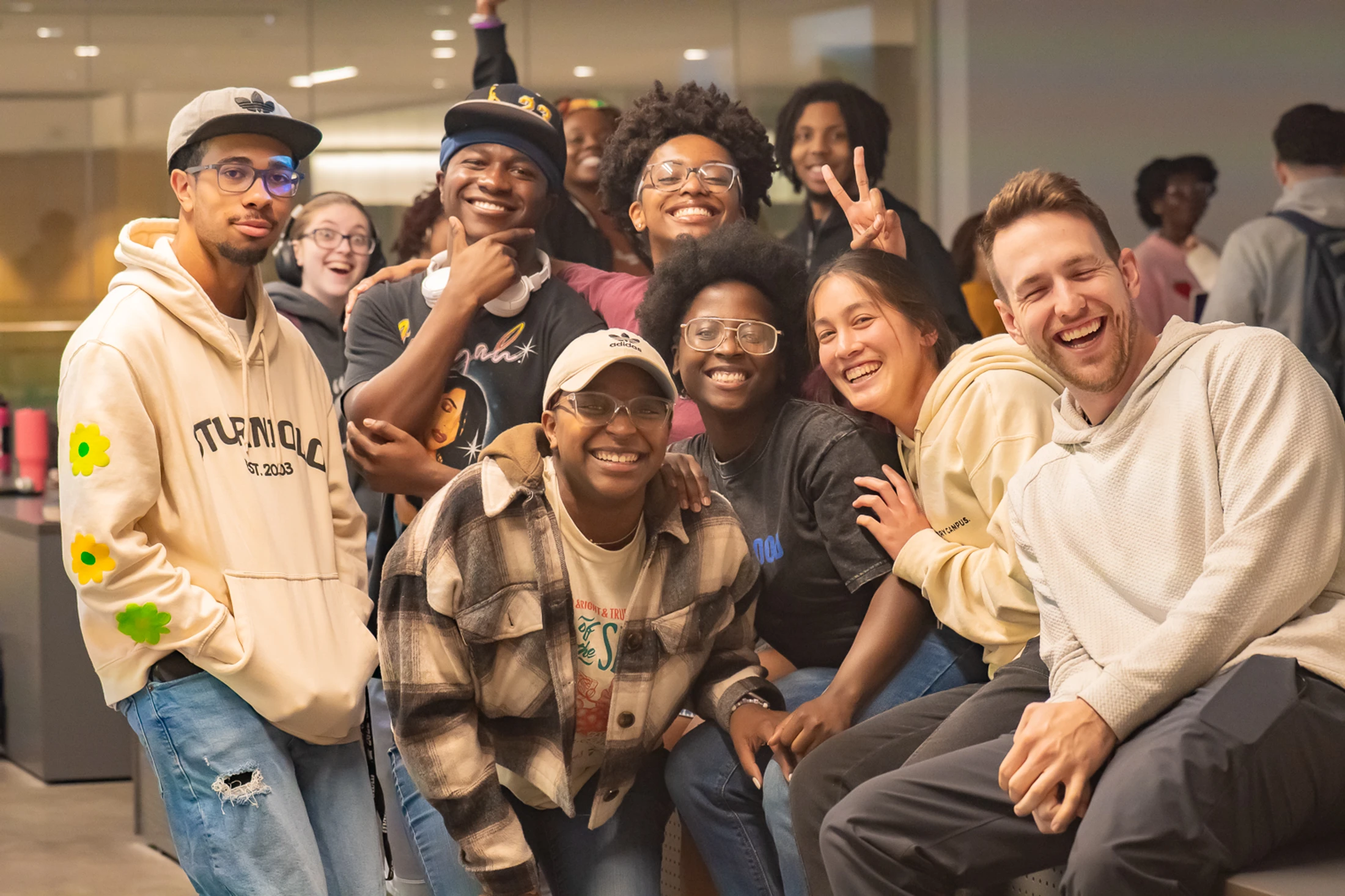 A diverse group of young adults smiling and laughing as they pose together for a group photo indoors. They are dressed in casual clothes and look happy.