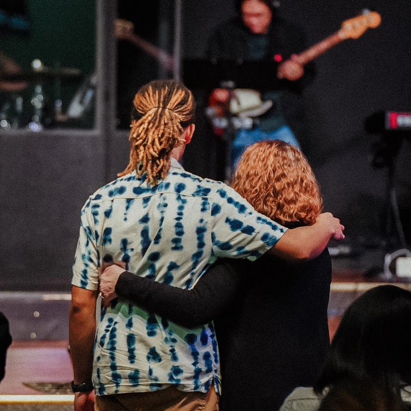 A man with blond dreadlocks and a woman with curly hair embrace from behind as they watch a musician playing a bass guitar on a dimly lit stage.