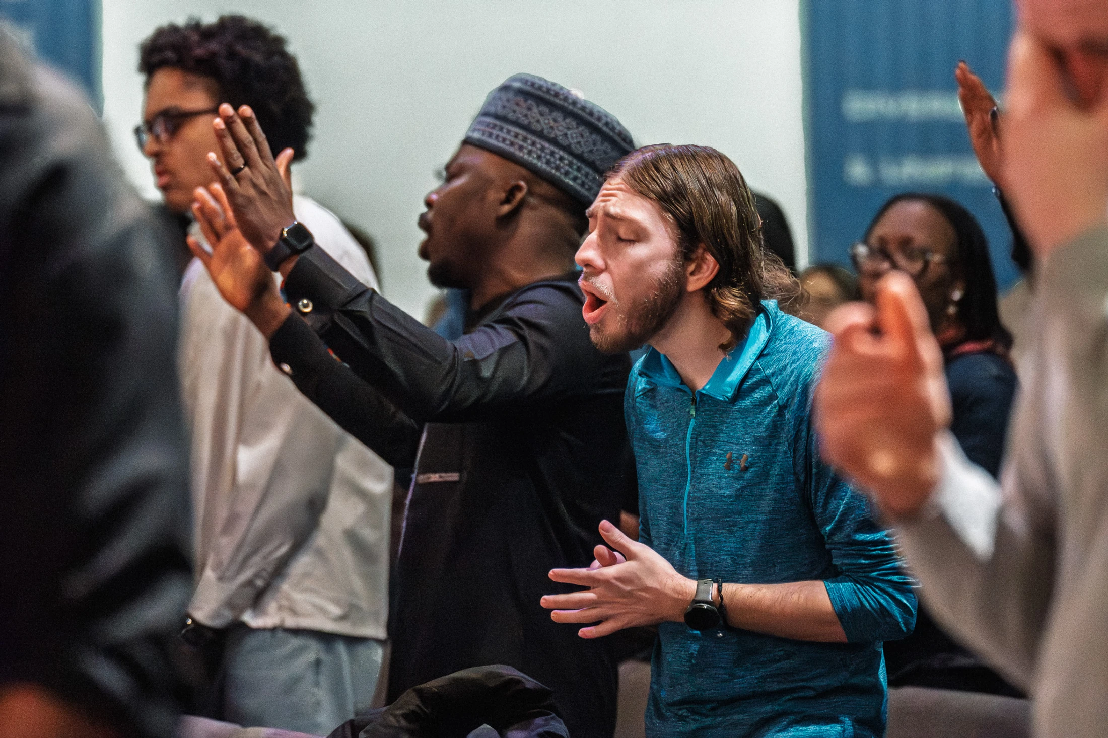 A diverse congregation worships together. In the foreground, a young man in a blue shirt sings with eyes closed, next to a man in a kufi hat with hands raised.