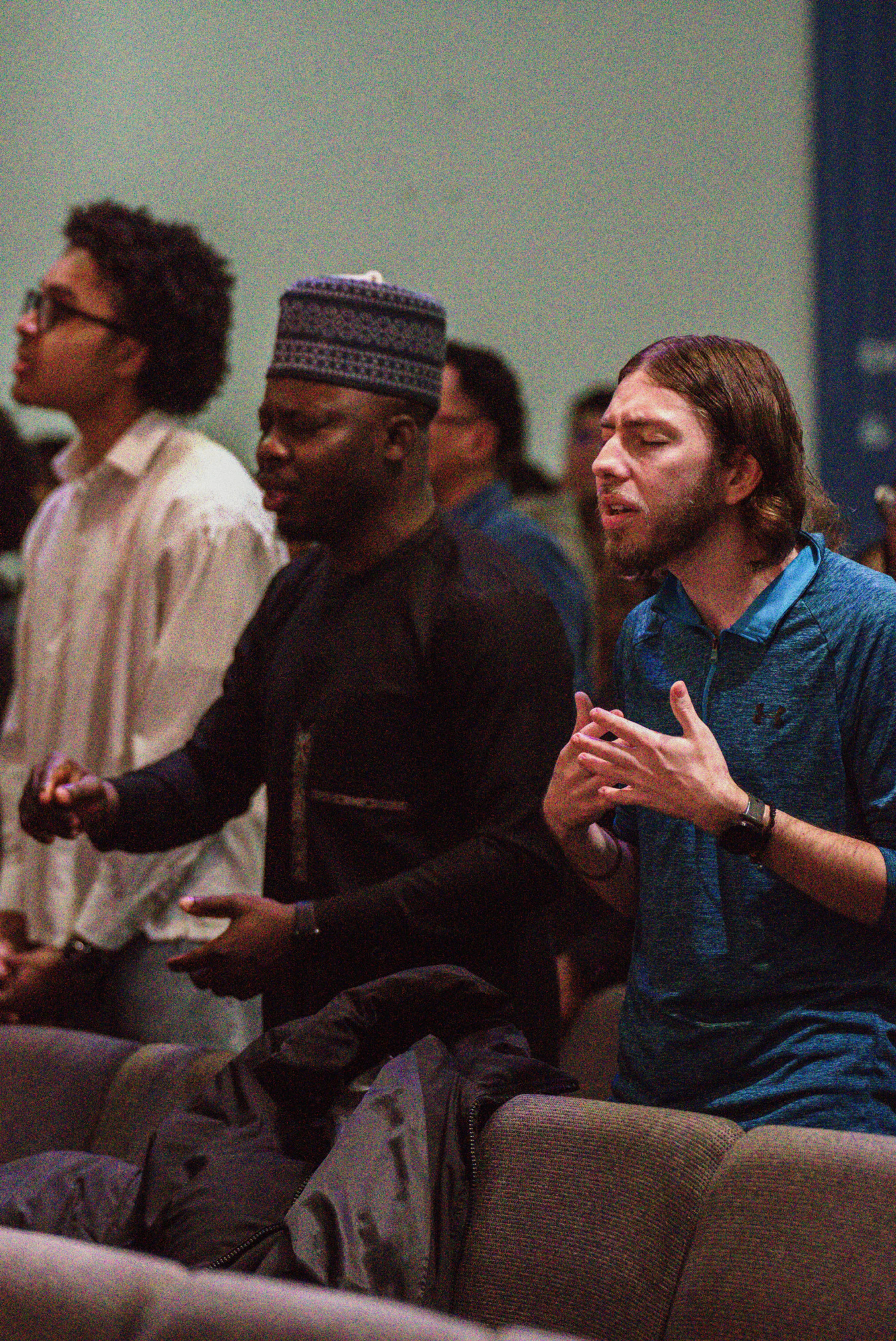 A diverse group of men stand together in a congregation, singing and praying with expressions of devotion during a service.