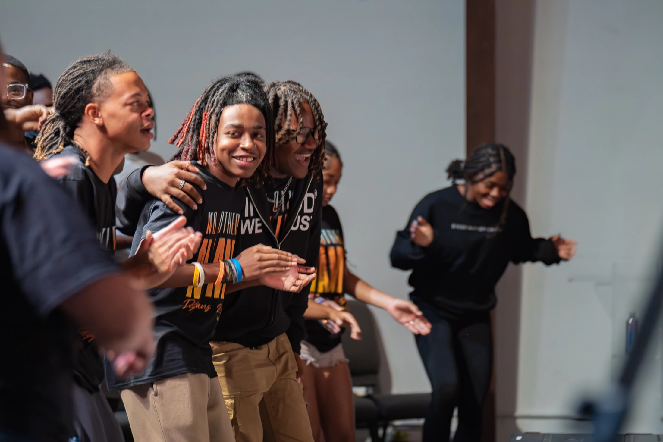 A group of young Black people joyfully clap and dance together indoors. One young man with locs smiles directly at the camera while others cheer him on.