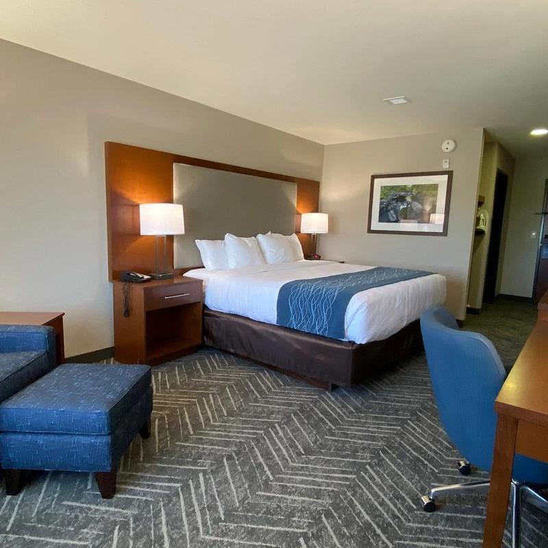 A hotel room featuring a king-sized bed with white linens, a blue armchair with a matching ottoman, and a wooden desk with a chair and lamp.