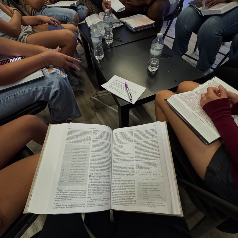 A first-person view of a Bible study, with an open Bible in the lap. A group of young people sits in a circle, also with Bibles, around a small table.