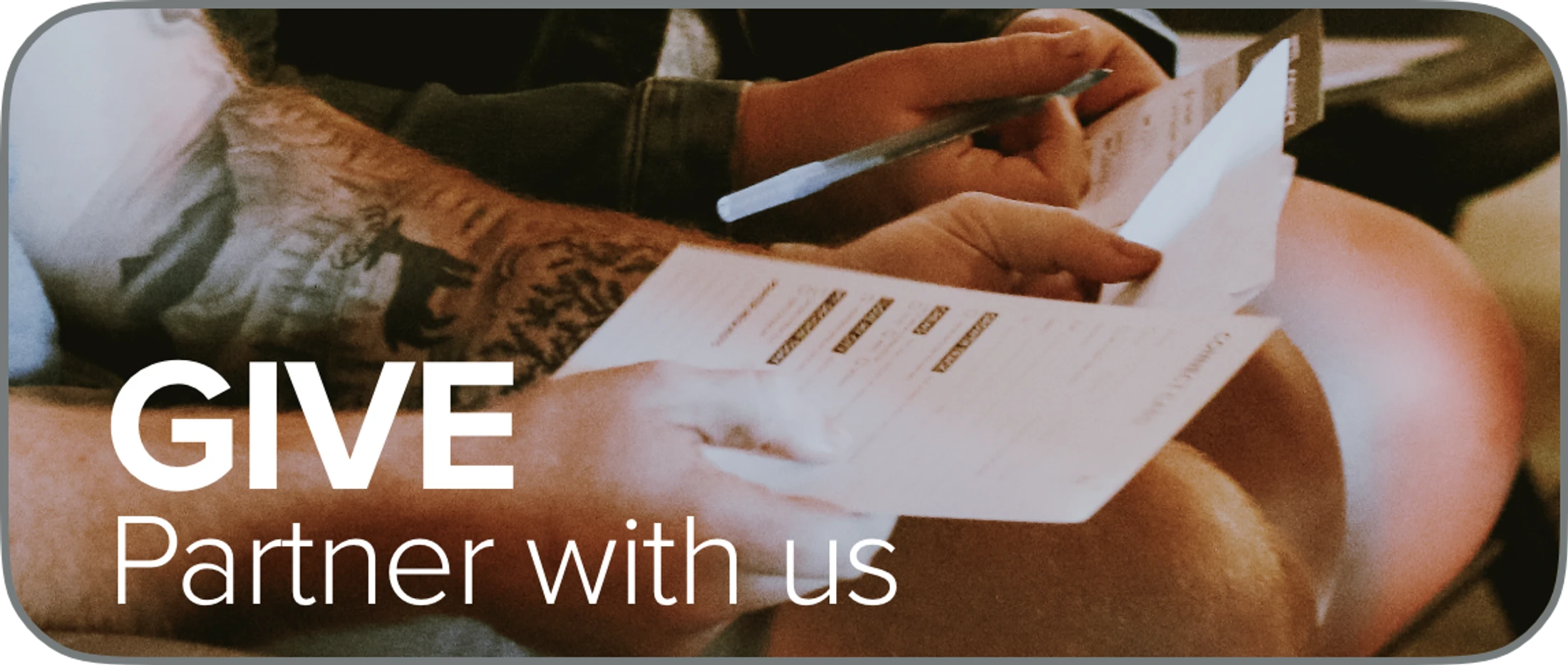 Close-up of a person with a tattooed arm holding a pen and filling out a donation form. Text on the image says "GIVE - Partner with us."