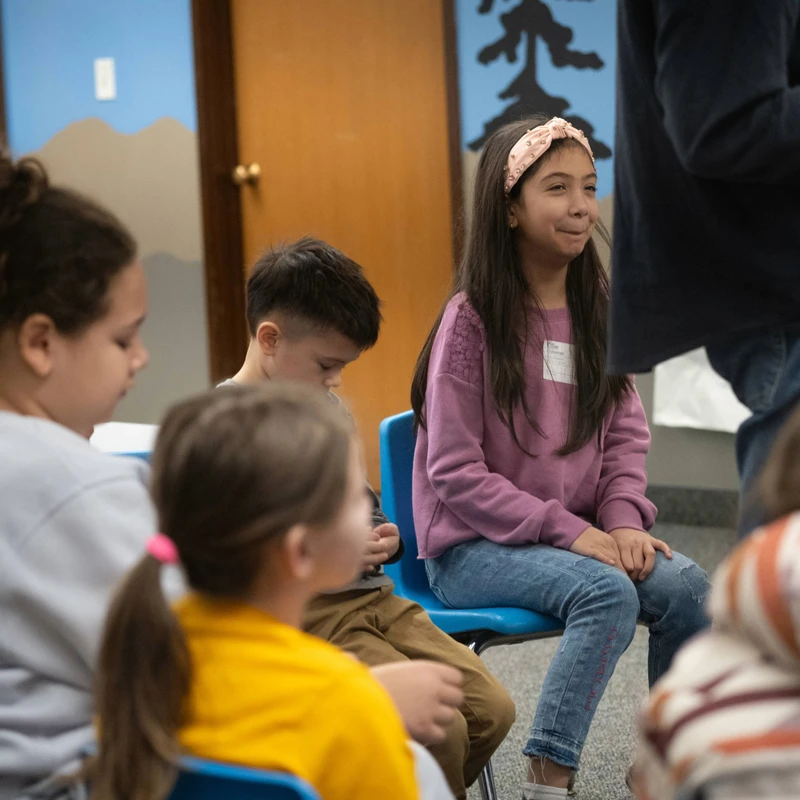 A group of young children sit in a classroom. A girl in the center, wearing a purple sweater and pink headband, smiles while looking at an adult.