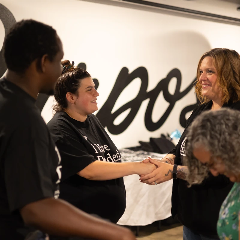 A pregnant woman in a black t-shirt shakes hands with another woman, both smiling. They are part of a small group of people at an indoor event.