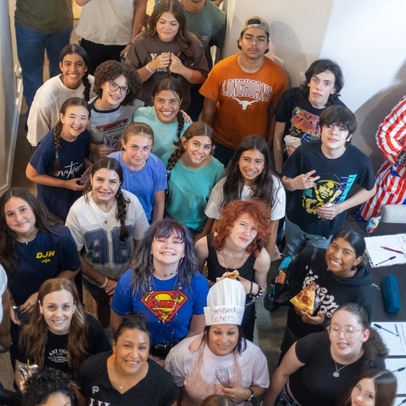 A high-angle photograph shows a large, diverse group of smiling teenagers and adults gathered together in a hallway, looking up at the camera.