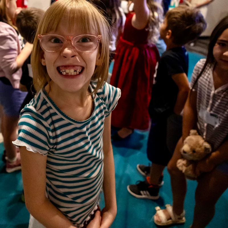 A young girl with blonde hair and pink glasses stands in a crowd, smiling widely at the camera and showing her teeth in a playful, silly grin.