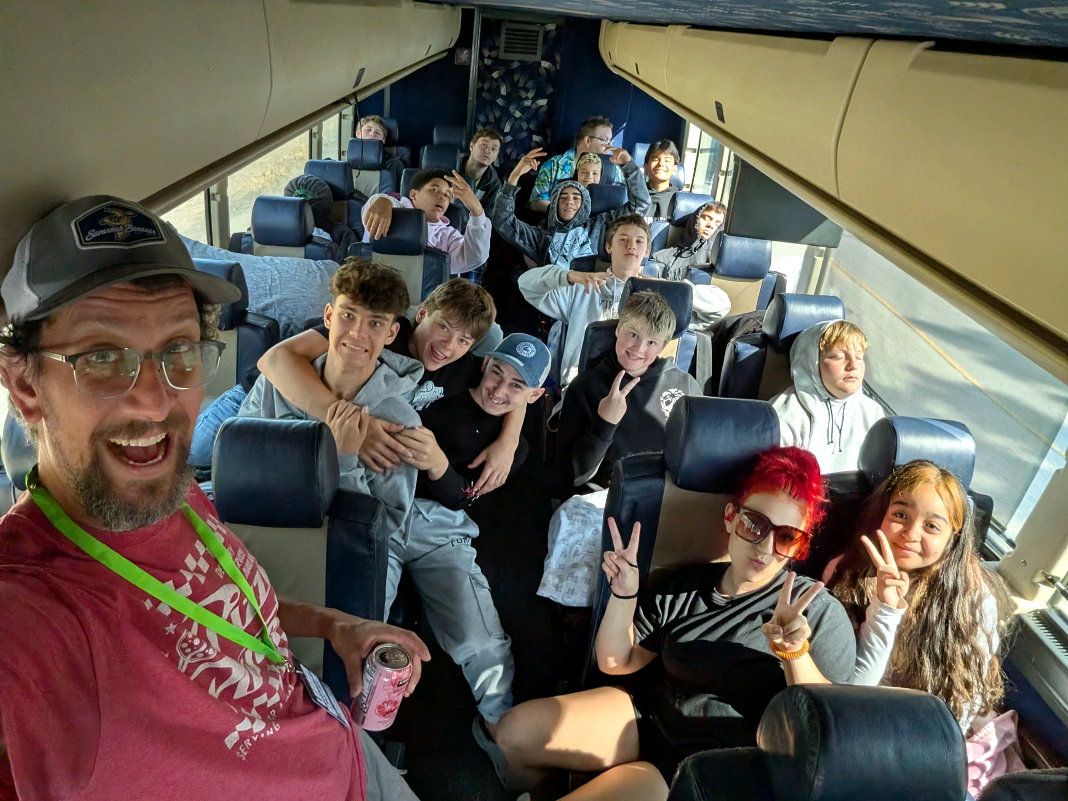 A man takes a wide-angle selfie with a large group of smiling teenagers posing behind him on a bus or train during a daytime trip.