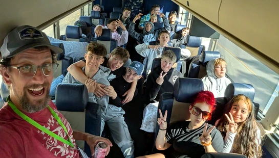 A man takes a wide-angle selfie with a large group of smiling teenagers posing behind him on a bus or train during a daytime trip.
