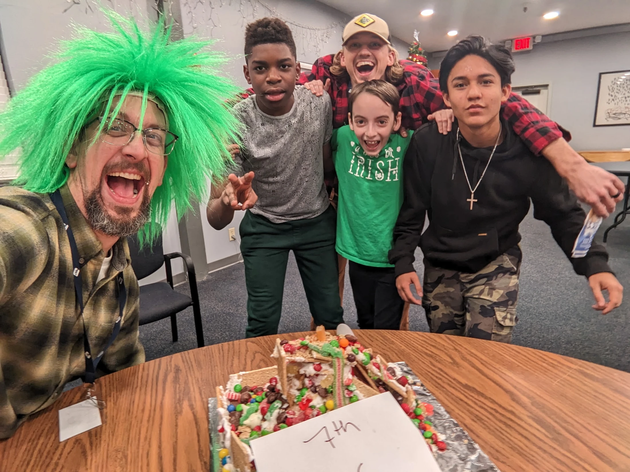 A man in a spiky green wig takes an excited selfie with four boys around a table with a messy, candy-covered gingerbread house on it.