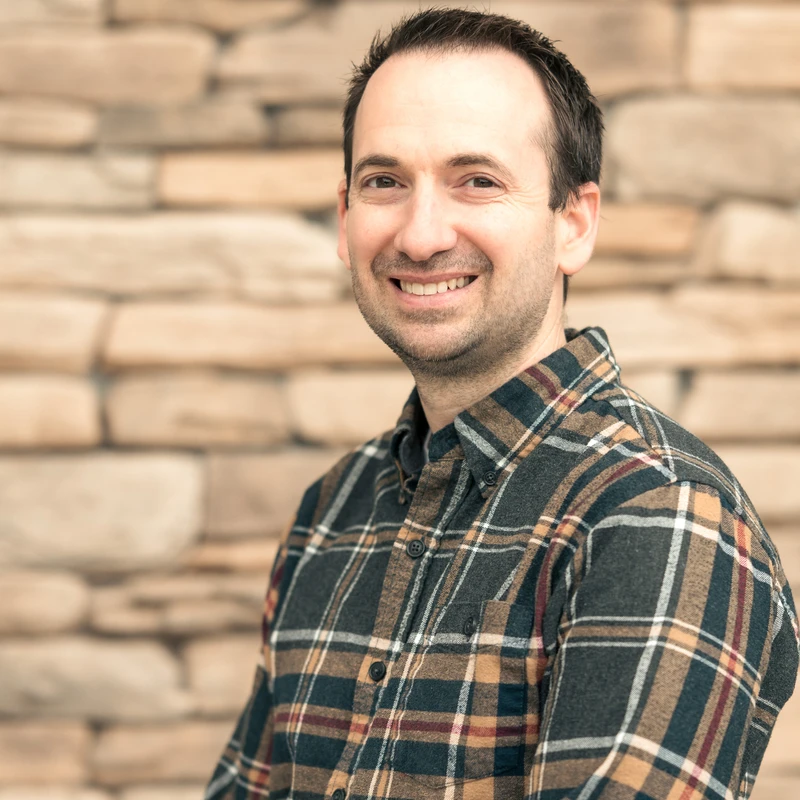 A man with short dark hair and a warm smile poses for a photo. He wears a dark plaid flannel shirt and stands in front of a blurred stone wall.