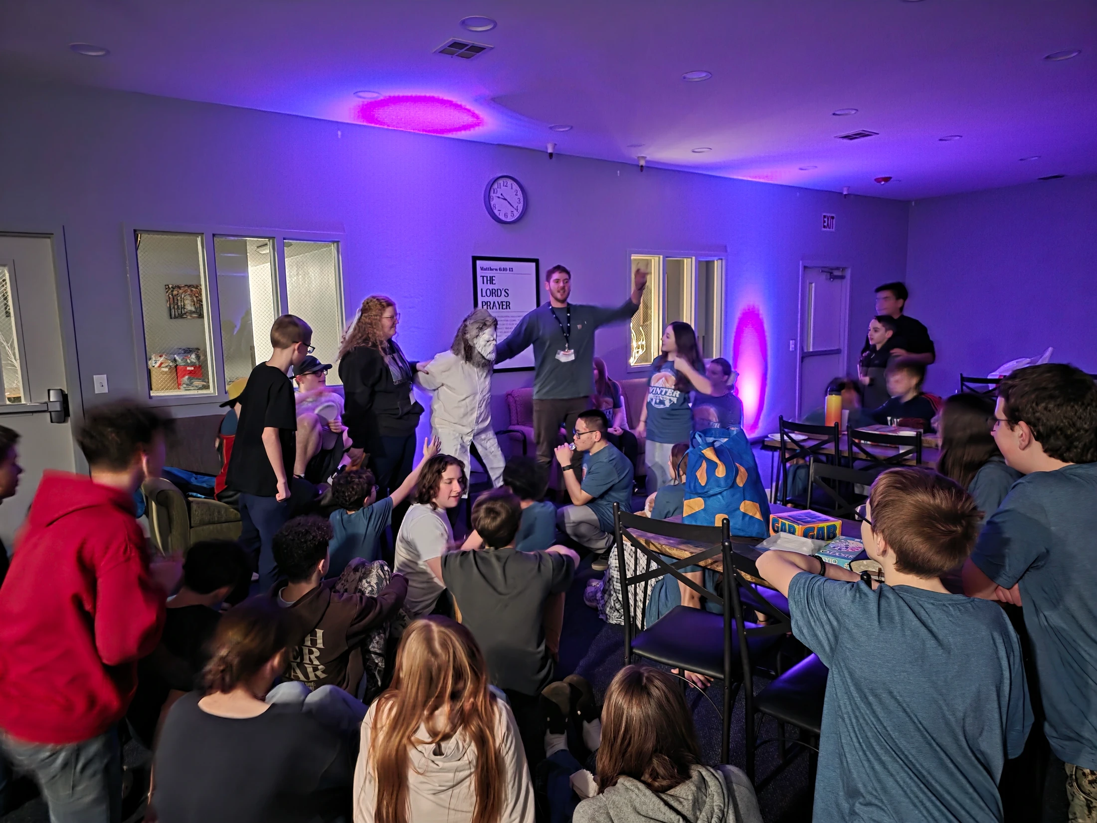 A large group of teenagers gathered in a purple-lit room, watching a man and another person in a white costume with a bearded mask at the front.