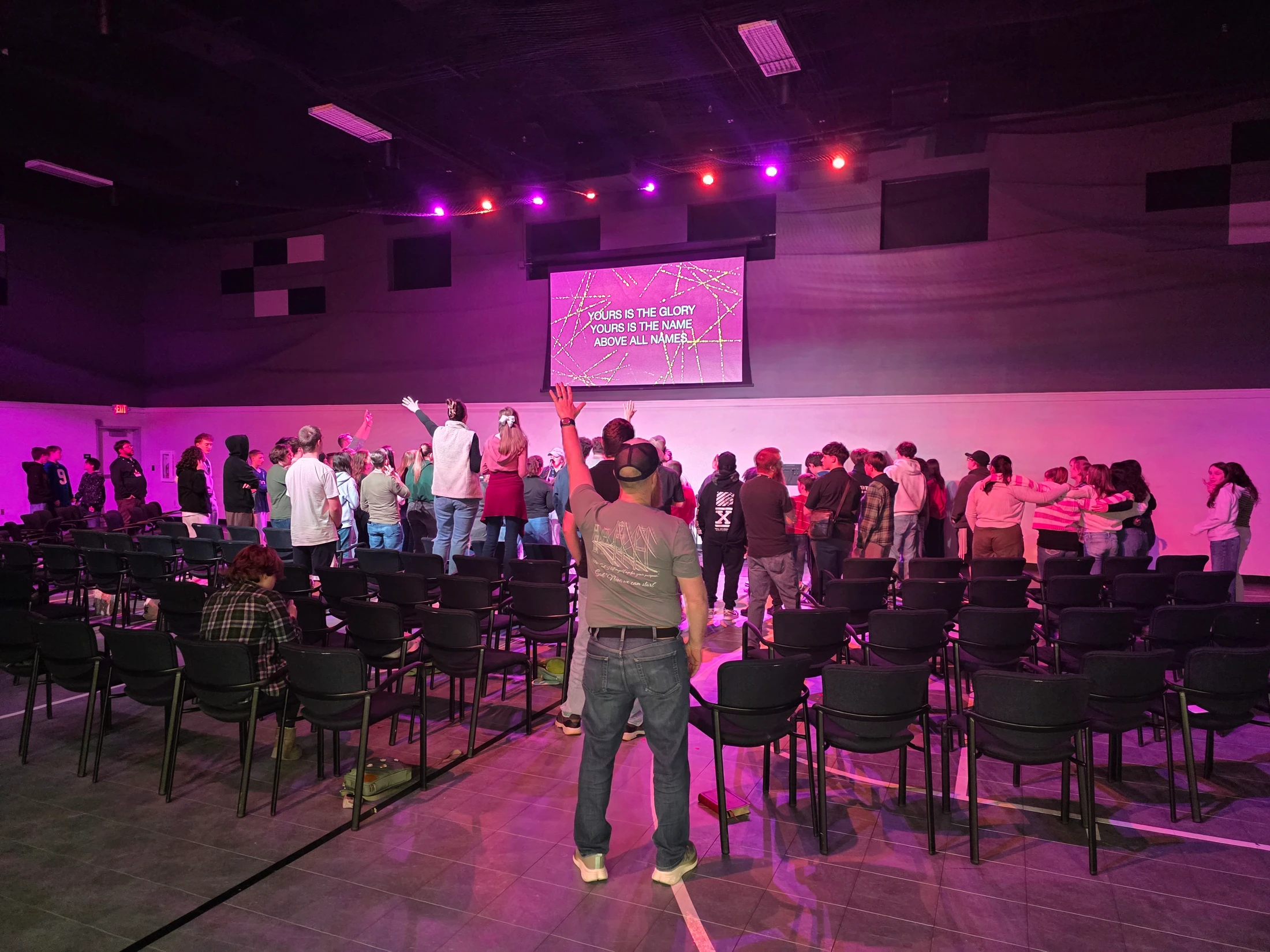 A large group of young people stand and worship in a dimly lit room with purple stage lights. Some raise their hands toward a screen with lyrics on it.