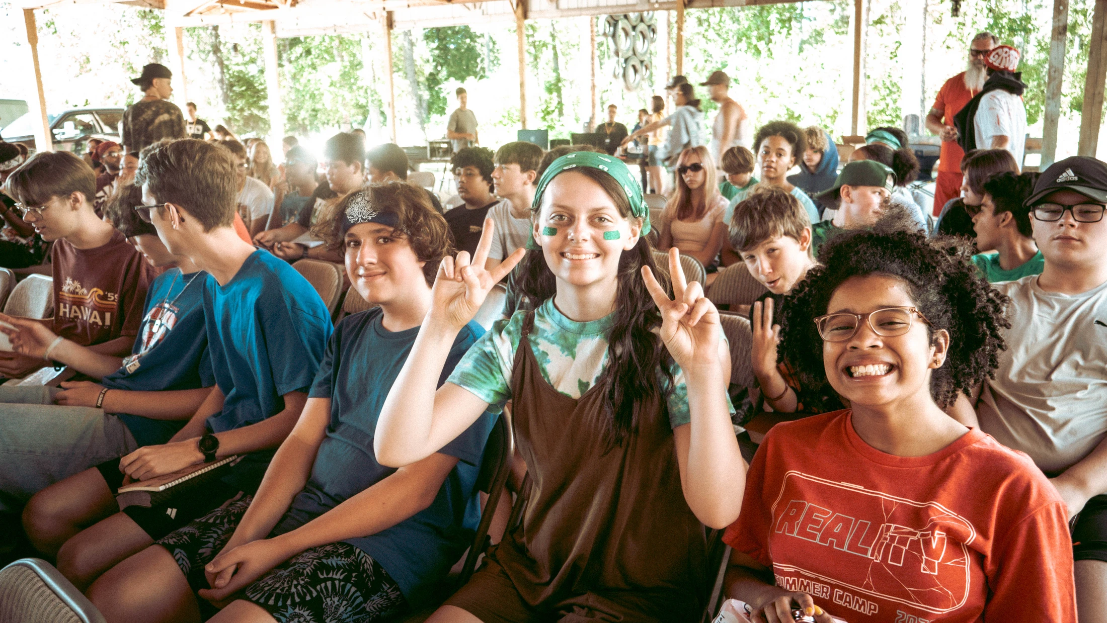 A large group of diverse teenagers sits together under an outdoor pavilion. Three young people in the foreground smile and pose happily for the camera.