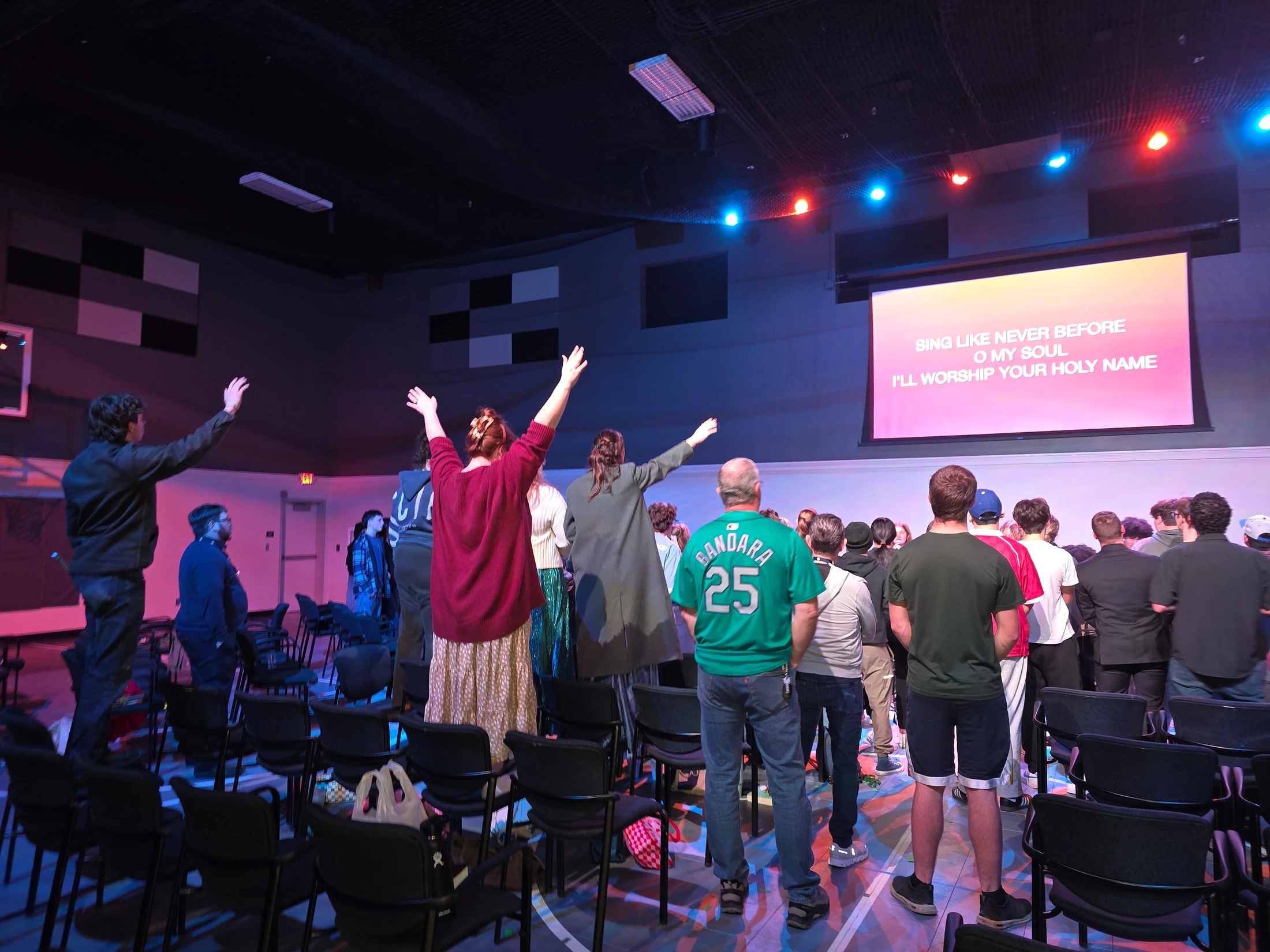 A diverse group of people stand in a dimly lit hall, facing a large screen with lyrics. Many have their hands raised in a gesture of worshipful singing.