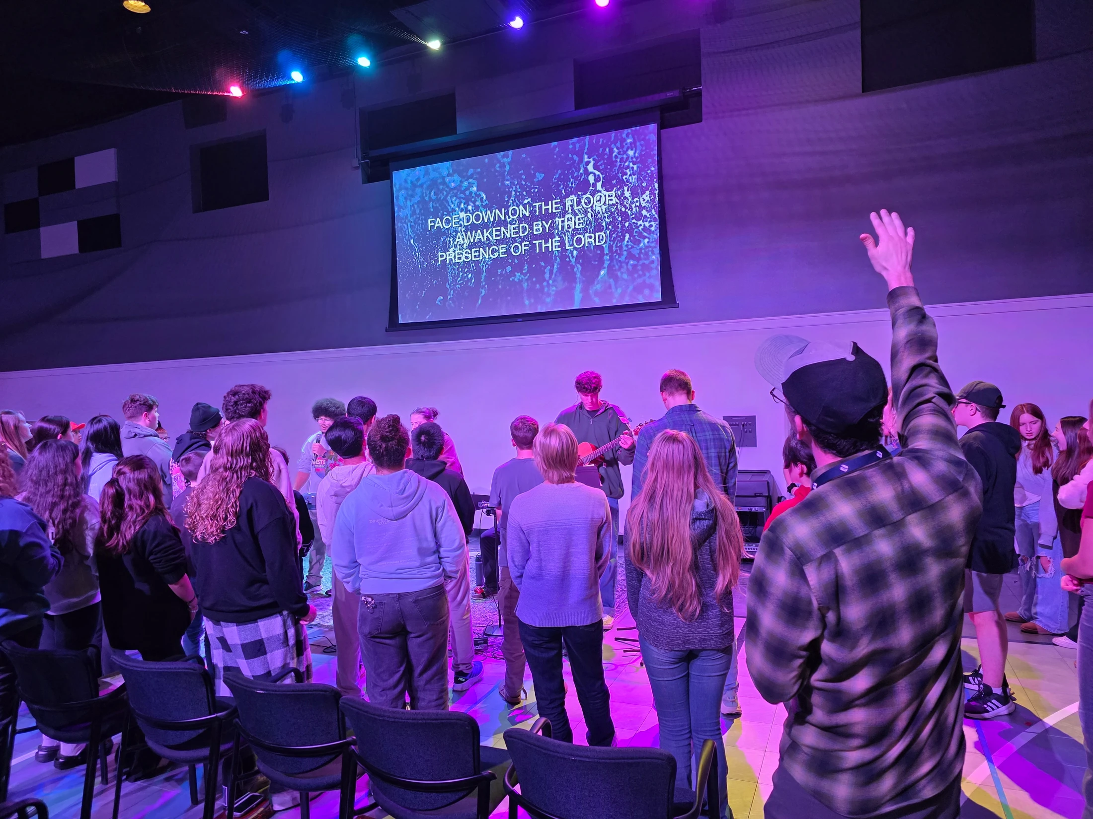 A crowd of young people stand and watch a musician play guitar on a stage lit with purple lights. A screen above shows lyrics for a worship song.
