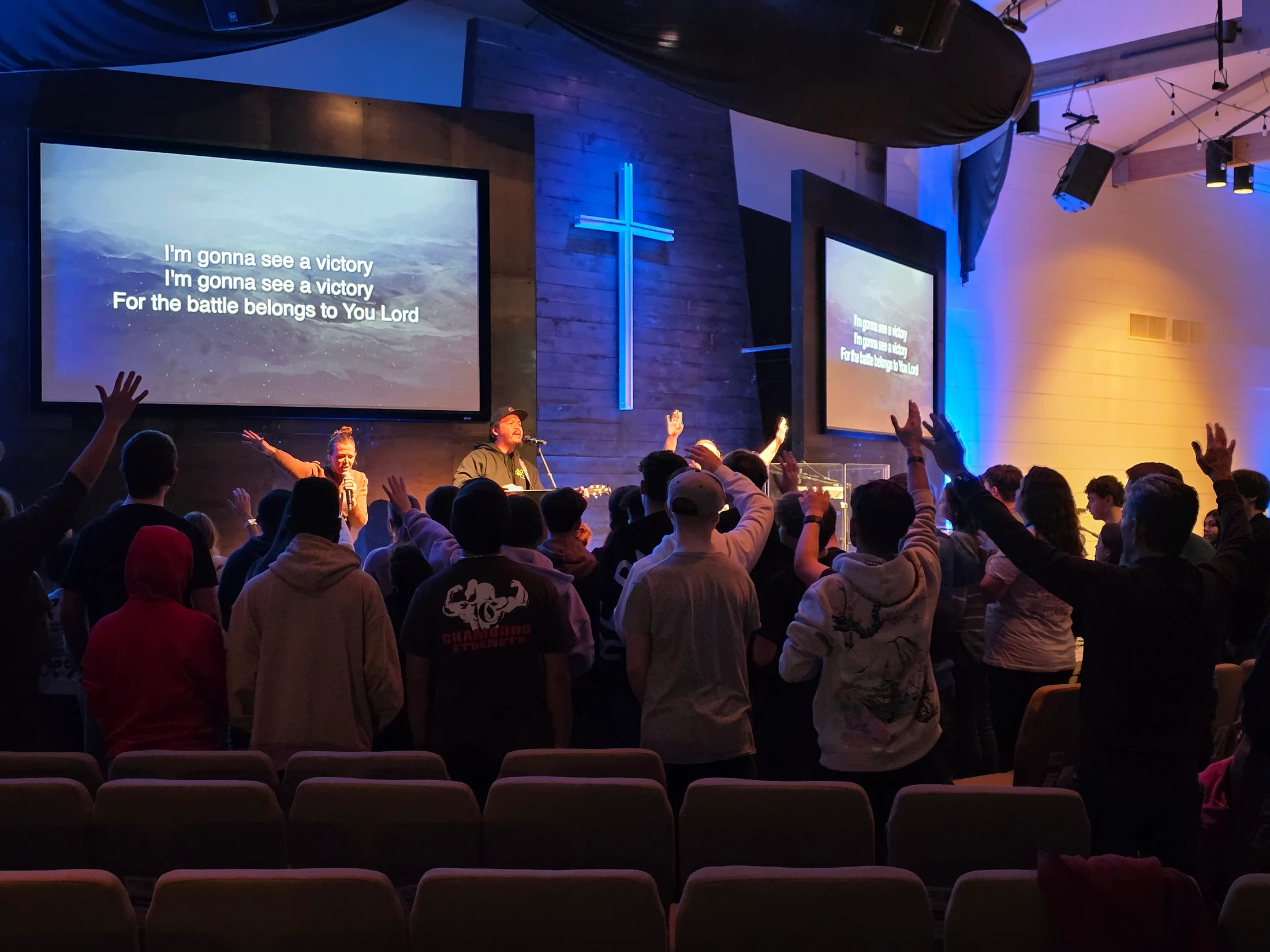 A view from behind a congregation with hands raised in praise during a modern church service, facing a stage with a band and screens showing lyrics.