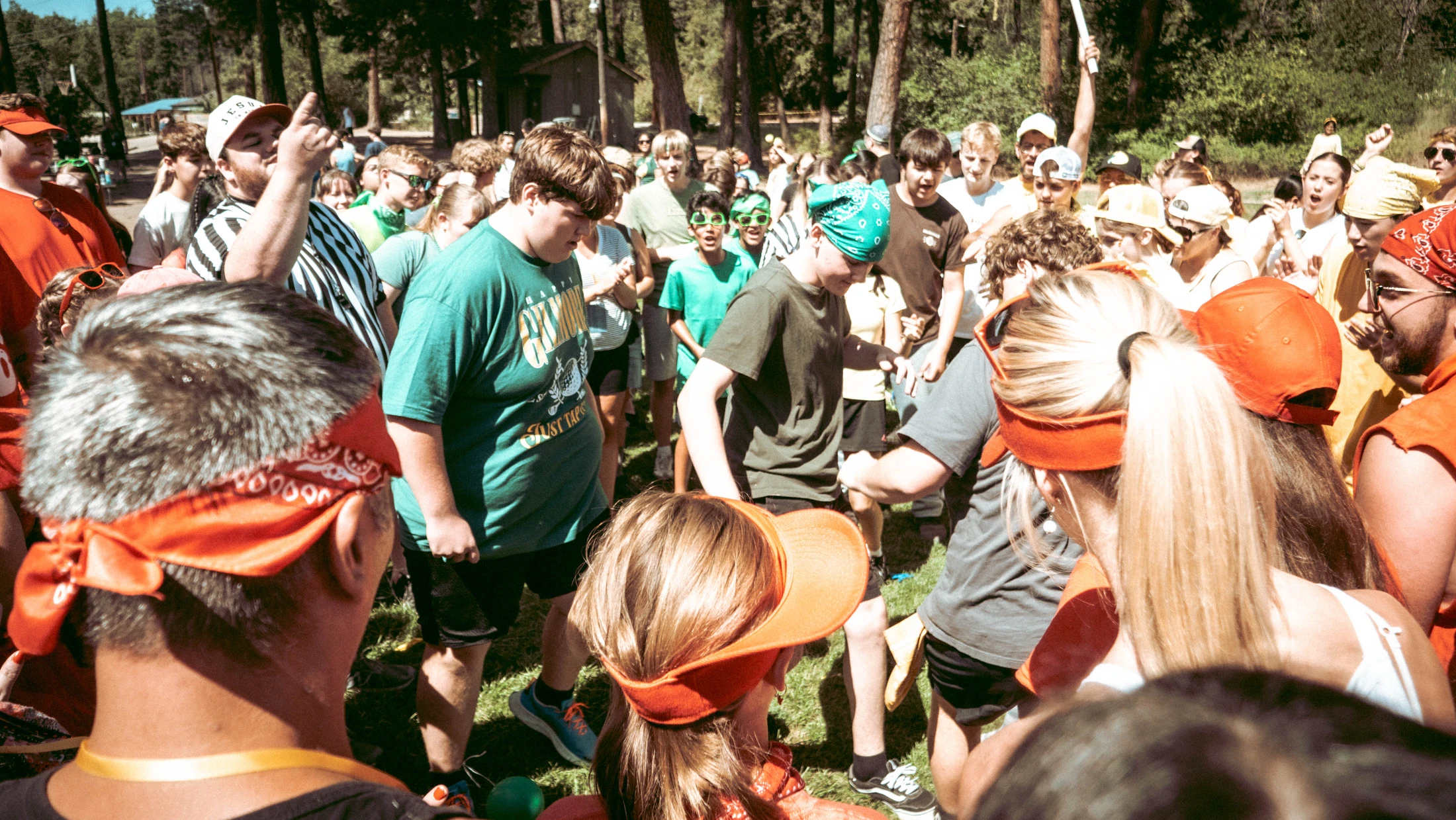 A large, diverse group of young people gathered outdoors in a circle, cheering during a summer camp activity on a sunny day in a wooded area.