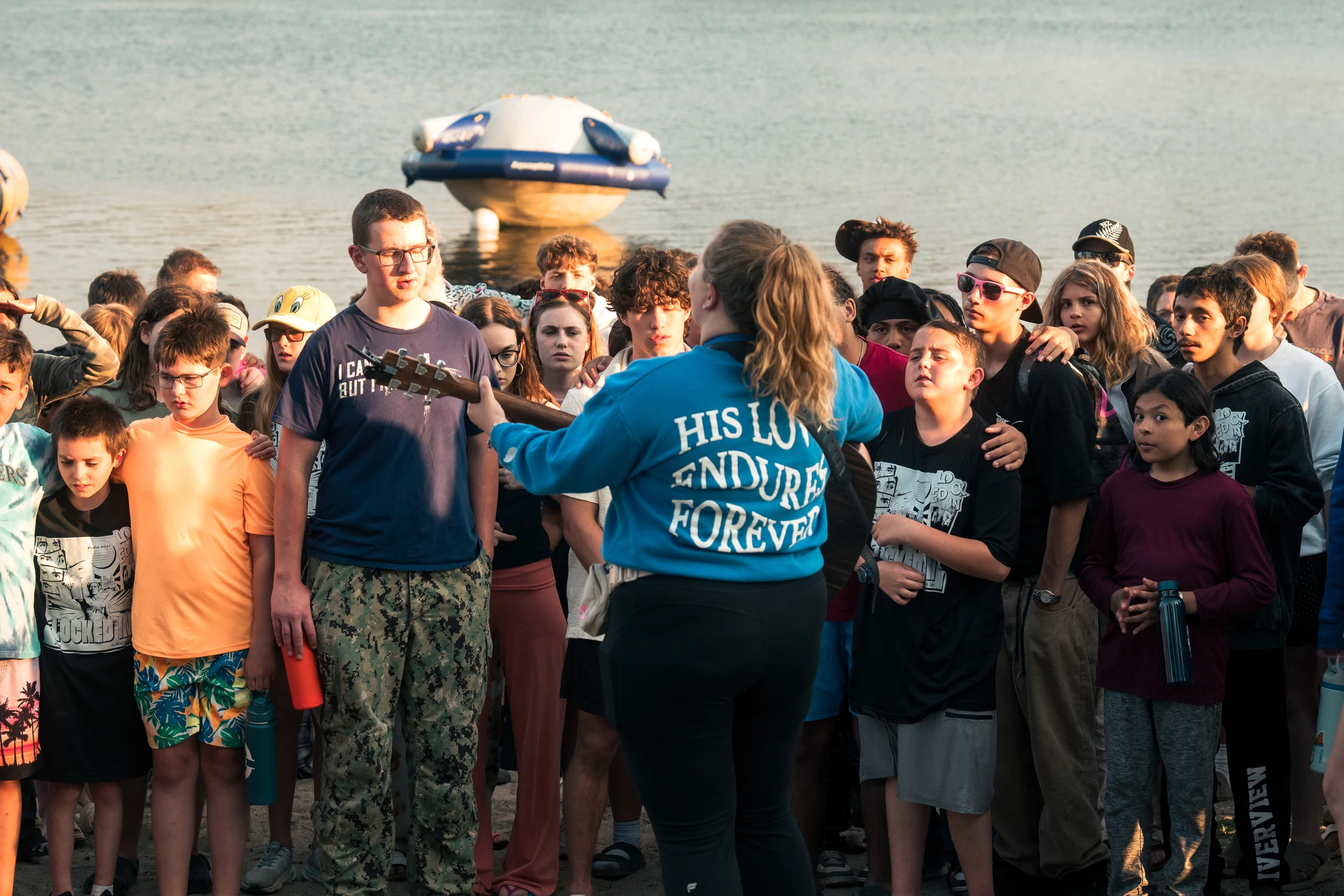 A group of young people stand on a shore, listening to a woman in a blue sweatshirt playing a guitar. The back of her shirt says 'HIS LOVE ENDURES FOREVER'.