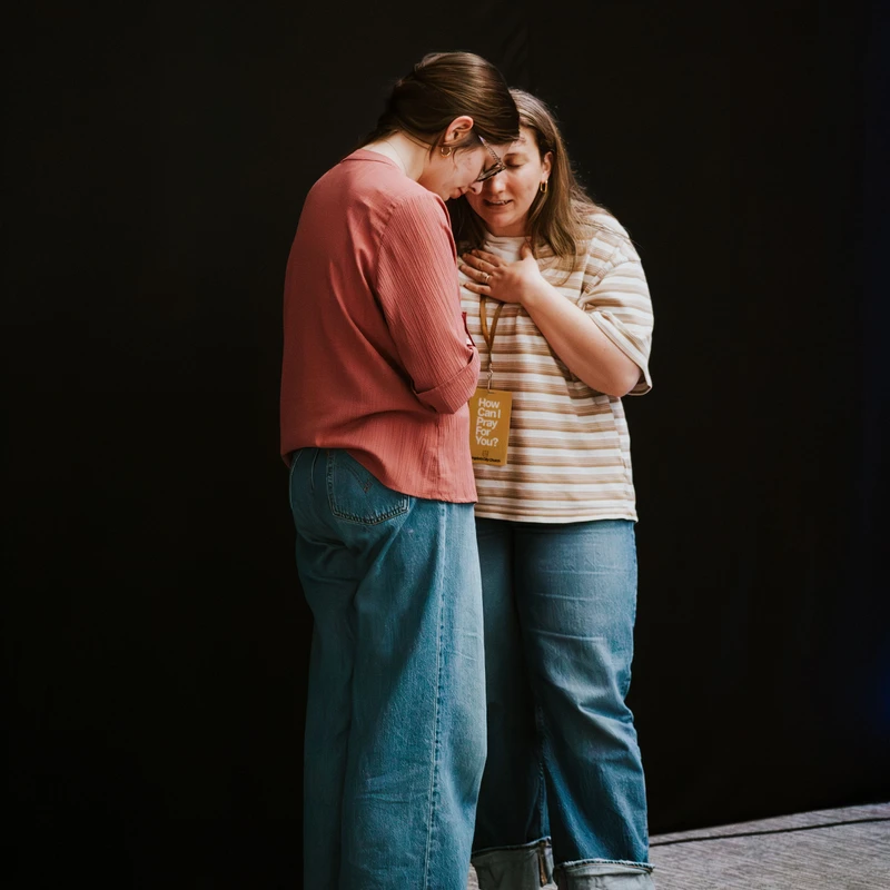 Two women stand close against a black backdrop, praying together. One has her hand on her chest and comforts the other, who has her head bowed.