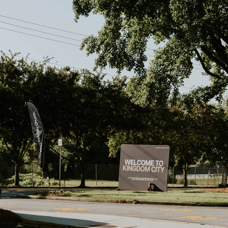 A gray welcome sign for Kingdom City Church stands on a grassy area next to a road. Large, leafy green trees are visible in the background under a sunny sky.