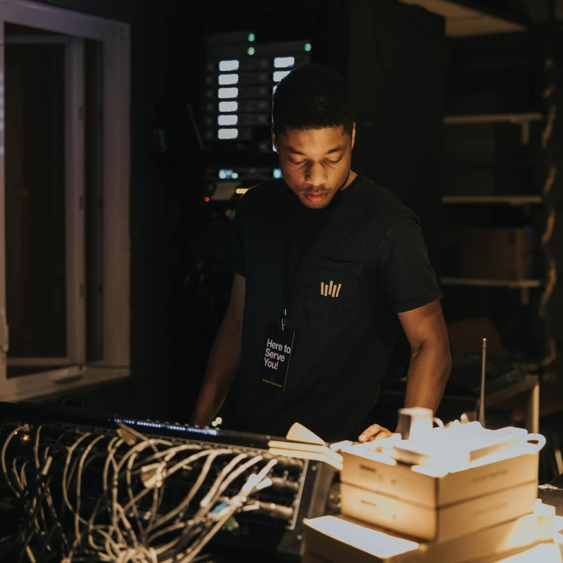 A young man with a lanyard that says 'Here to Serve You' stands in a dimly lit room, focused on a complex sound mixing board covered in cables.