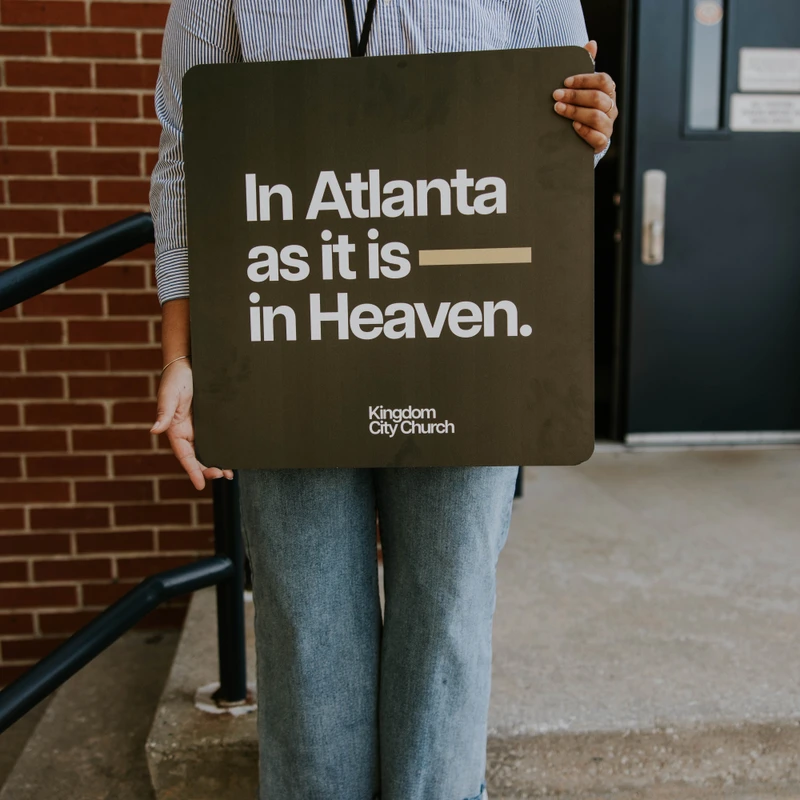 A smiling woman in a striped shirt and red shoes holds a sign that reads 'In Atlanta as it is in Heaven' from Kingdom City Church.