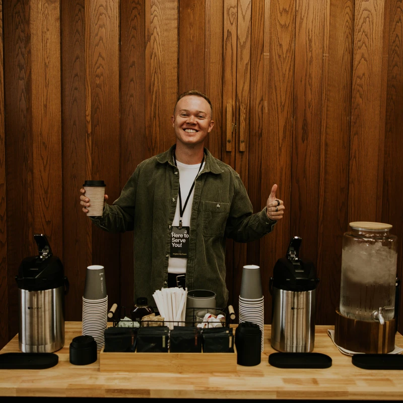 A smiling young man stands behind a coffee and beverage station, holding a cup and giving a thumbs up. He wears a badge that says "Here to Serve You!"