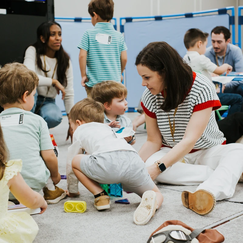 A group of young children and several adults sit on a grey carpeted floor, interacting and playing with toys in a classroom or childcare setting.