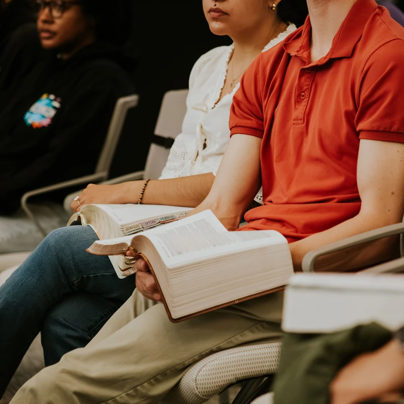 A diverse group of people sits in a row of chairs, listening. A man in a red shirt and a woman next to him hold thick, open books that appear to be Bibles.