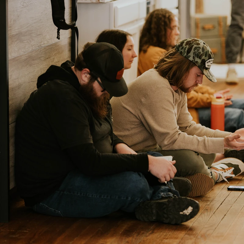 A group of people sitting on a hardwood floor, leaning against a wall. A man in a camo hat has his hands open, as if in prayer or discussion.