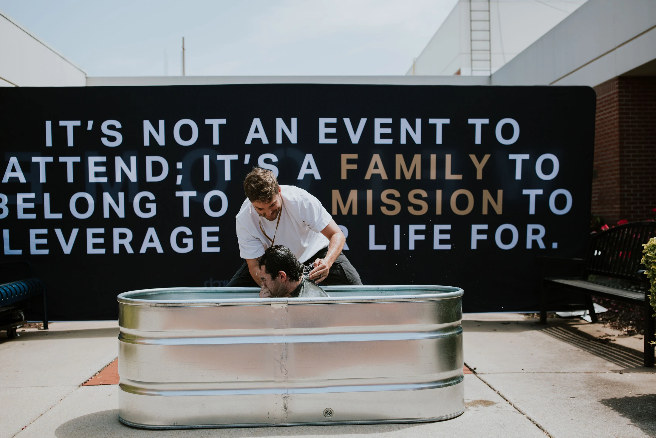 A pastor baptizes a man by immersion in a metal stock tank outdoors. Behind them is a large black banner with white and gold text about community.