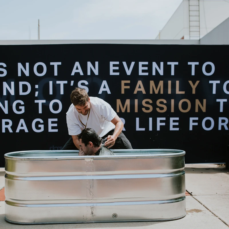 A pastor baptizes a man by immersion in a metal stock tank outdoors. Behind them is a large black banner with white and gold text about community.