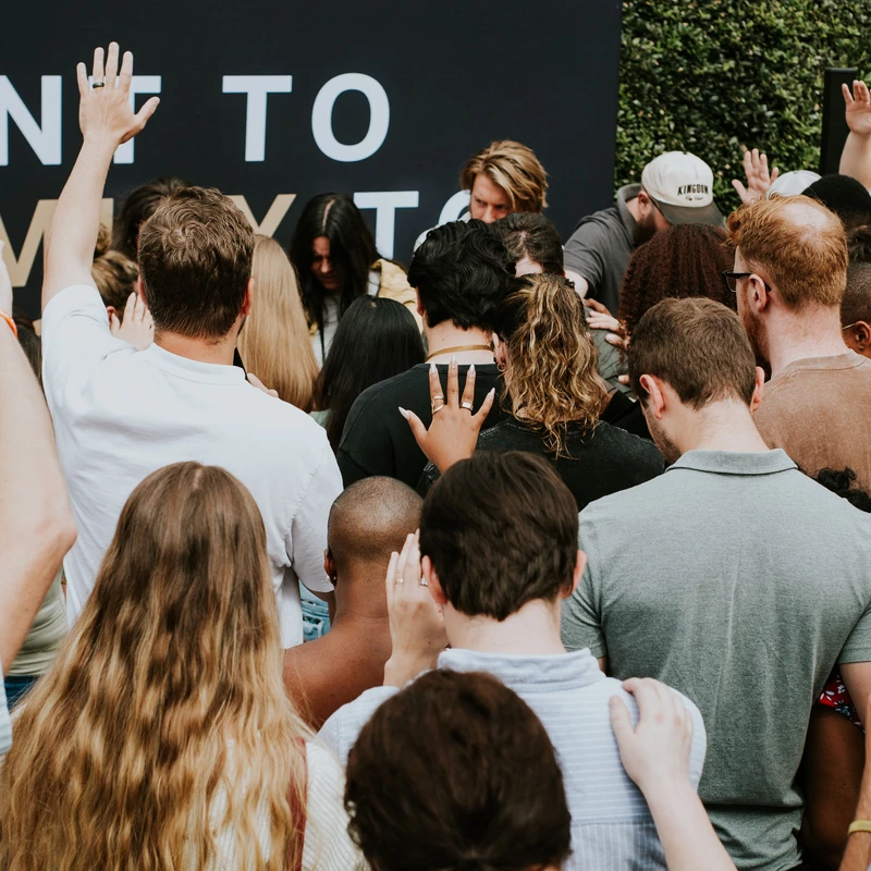 A large group of people seen from behind, huddled together with hands raised and on each other's shoulders in prayer in front of a large sign.