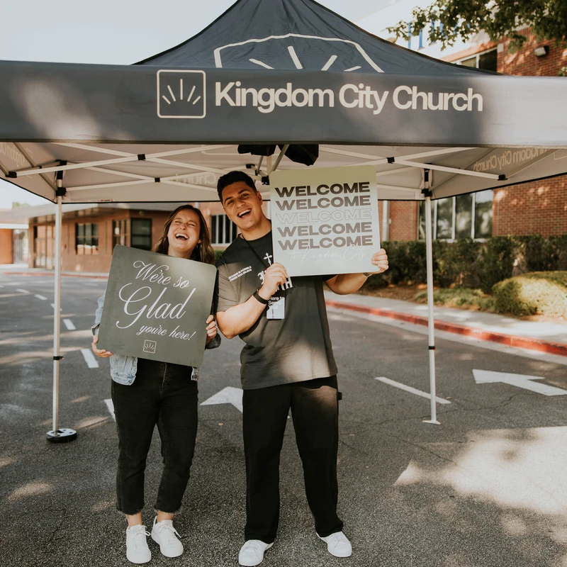 A man and a woman stand smiling under a Kingdom City Church tent in a parking lot, holding welcome signs for visitors.
