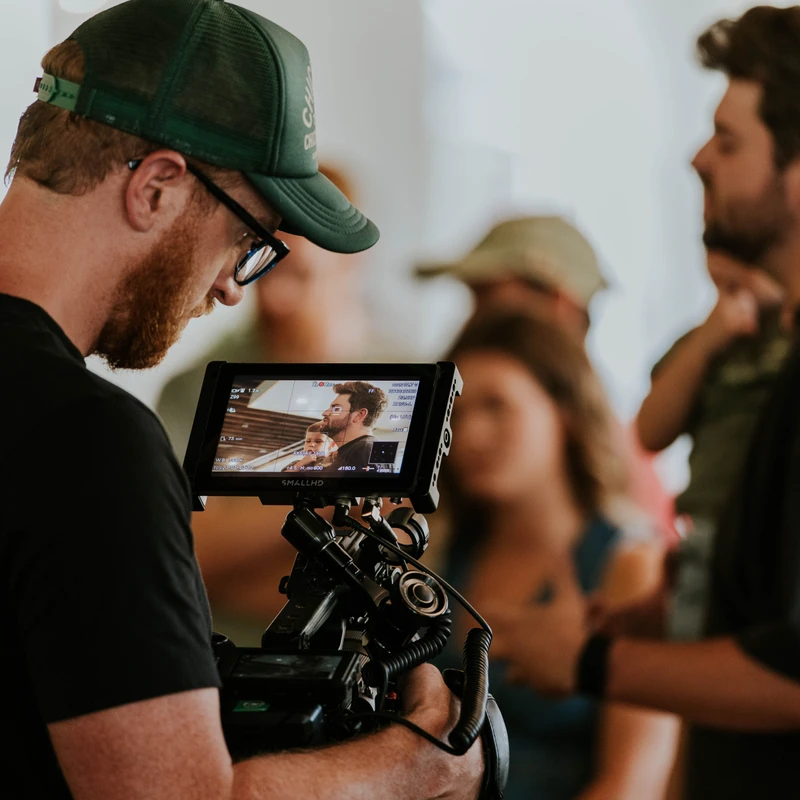 A cameraman with a red beard and green cap looks down at his camera's monitor, which shows the scene he is filming. People are blurred in the background.