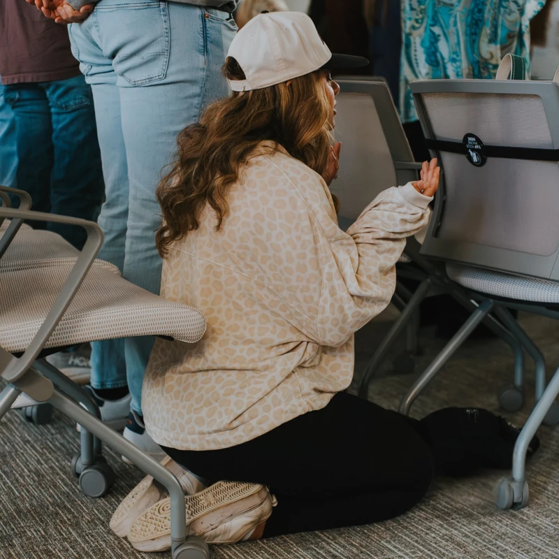 A woman with long hair in a backward cap and leopard-print sweatshirt kneels on the carpeted floor to pray in front of an empty chair, surrounded by others.