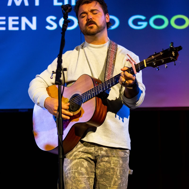 A man stands on a stage with his eyes closed, singing into a microphone and playing an acoustic guitar. Behind him, a blue screen displays song lyrics.