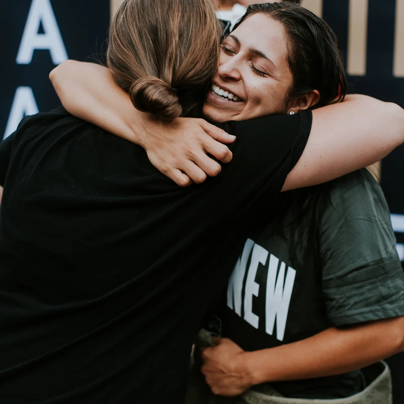 A woman with wet hair and a green t-shirt smiles with her eyes closed while hugging another woman, wrapped in a green towel after her baptism.