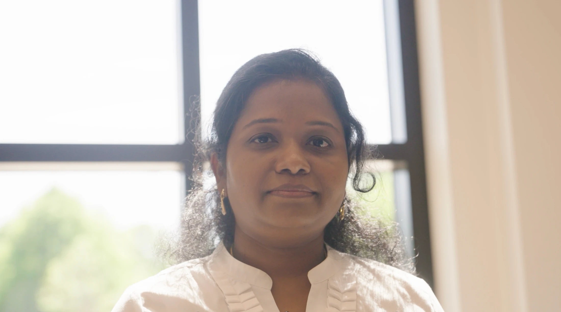 A headshot of a woman with dark curly hair and brown eyes, wearing a white collared shirt. She is looking at the camera, backlit by a bright window.