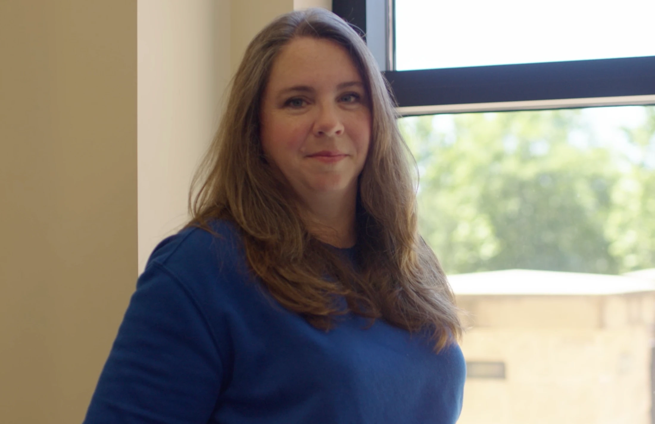 A woman with long brown hair and a blue sweatshirt smiles gently at the camera while standing indoors next to a window.