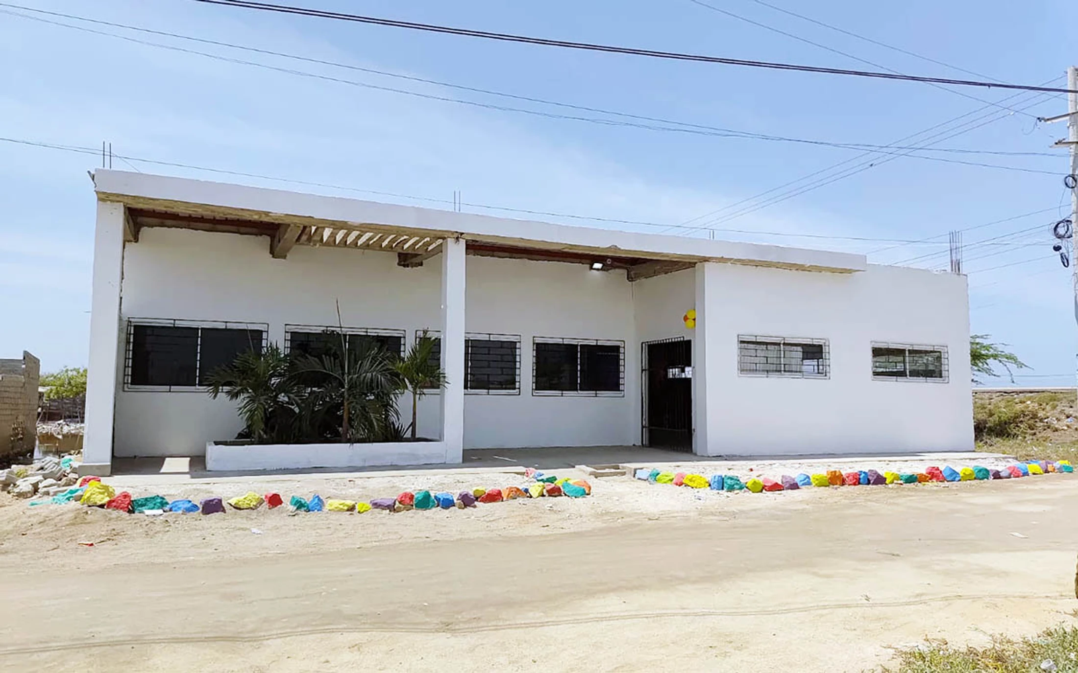 A one-story white building, possibly under construction, sits beside a dirt road. A line of brightly painted rocks decorates the front of the property.