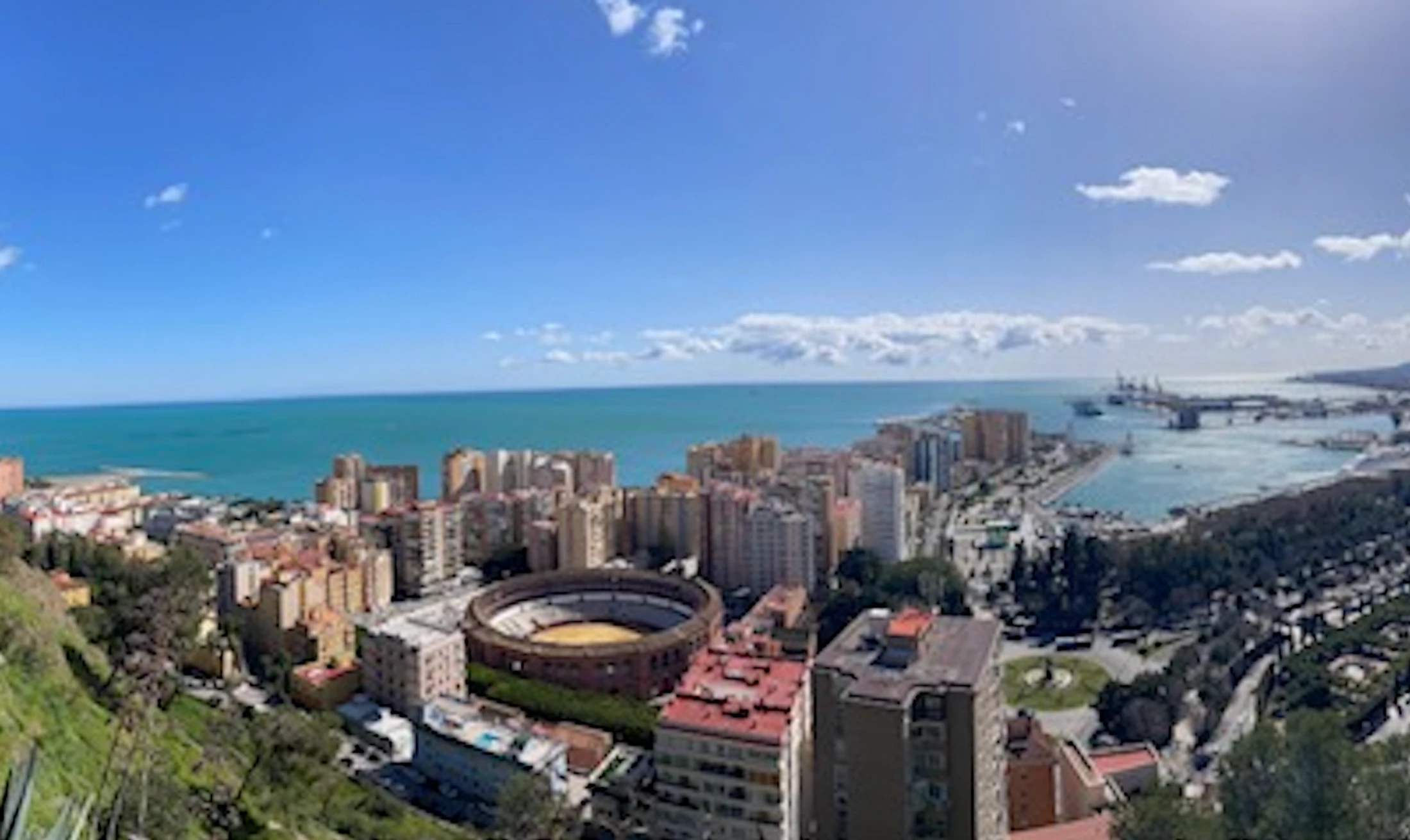 A sunny, elevated panoramic view of Málaga, Spain. The image shows the city buildings, the La Malagueta bullring, the port, and the turquoise Mediterranean Sea.