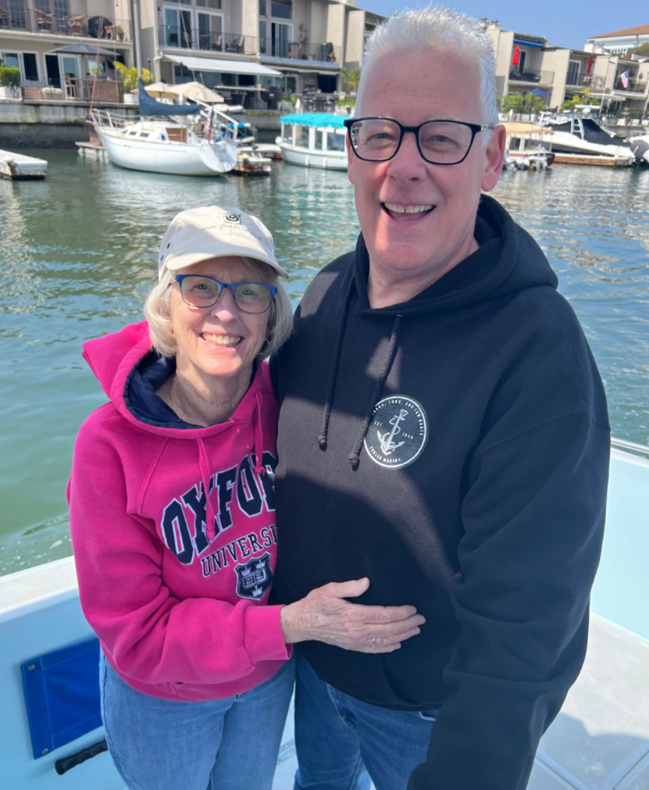 A smiling older man and woman pose together on a boat. The woman wears a pink Oxford hoodie, and the man wears a black hoodie. A marina is in the background.