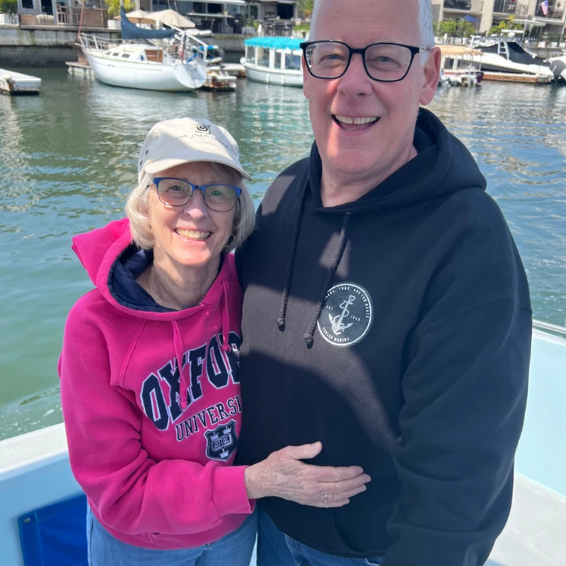 A smiling older man and woman pose together on a boat. The woman wears a pink Oxford hoodie, and the man wears a black hoodie. A marina is in the background.