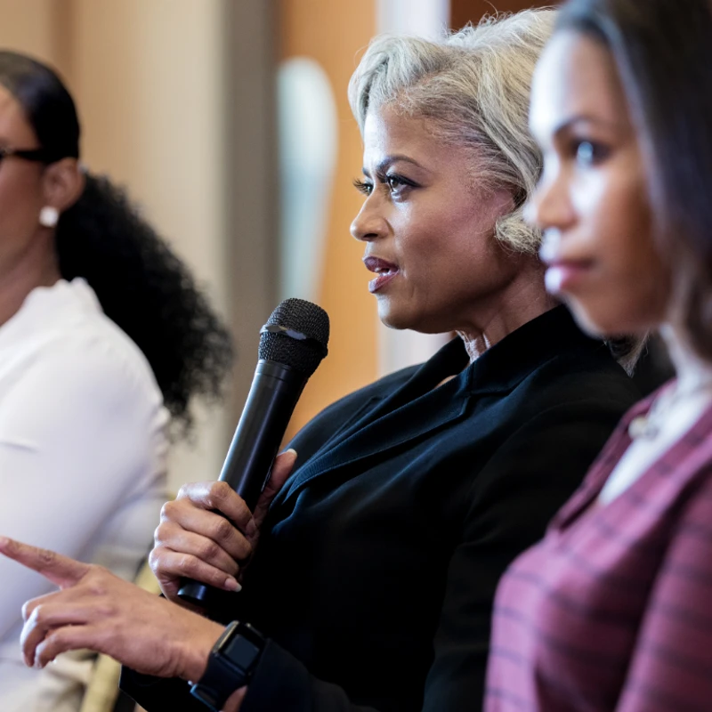 A woman with styled gray hair speaks into a microphone while gesturing. Two other women are seated beside her, one in focus and one out of focus.
