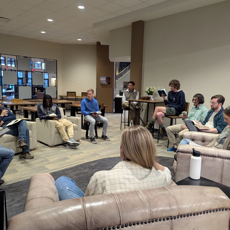 A diverse group of young adults sit in a circle in a common area, holding books and attentively listening during a bible study or discussion group.